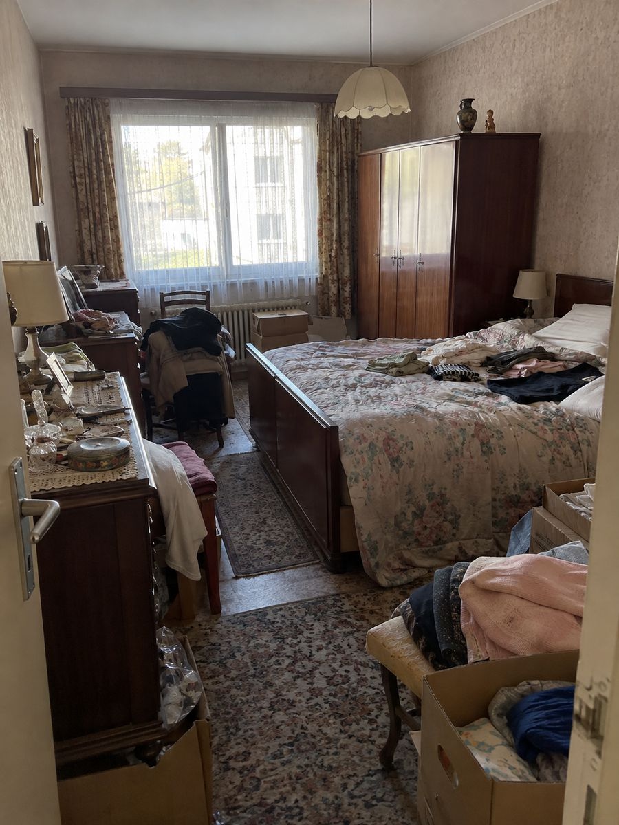 Chambre d’appartement des années 1970 d’une personne âgée avec garde-robe, lit, commode, boîtes et piles de vêtements et d’effets personnels