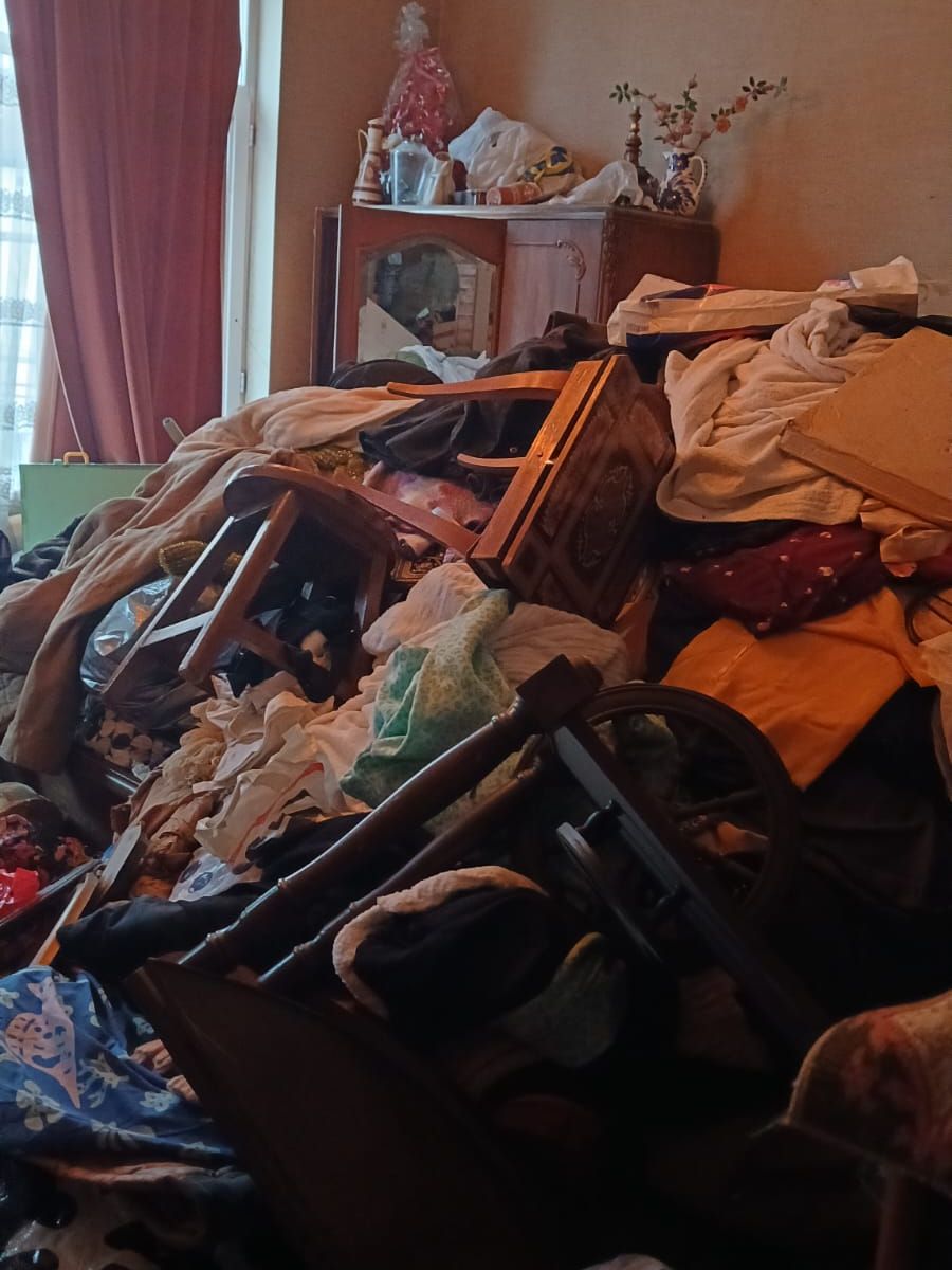 Salon d’une maison ancienne à Charleroi encombré de linge, de chaises en bois renversées et d’objets entassés devant un buffet vitré
