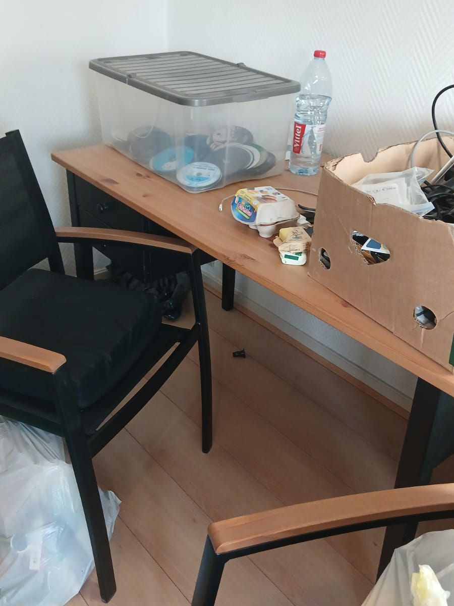 Coin bureau à Mons avec table en pin, chaises noires, boîte de CD, carton de câbles et sacs au sol